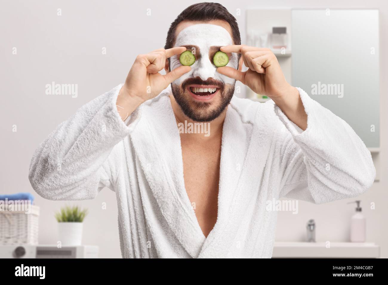 Man in a bathrobe with a face mask holding cucumbers on eyes in a bathroom at home Stock Photo