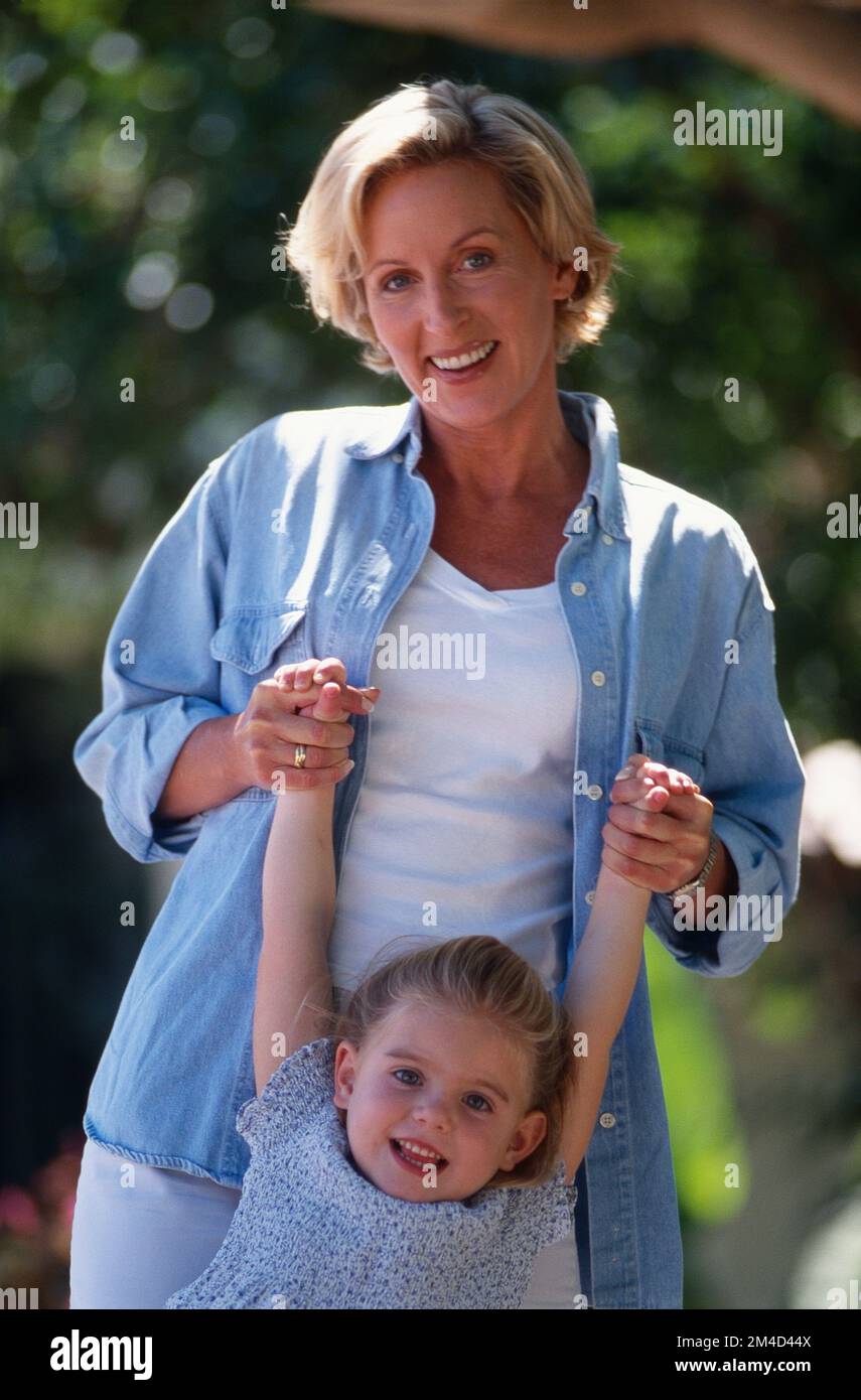 Young blonde headed mother standing behind her little girl holding her hands up in the air both smiling at the camera Stock Photo