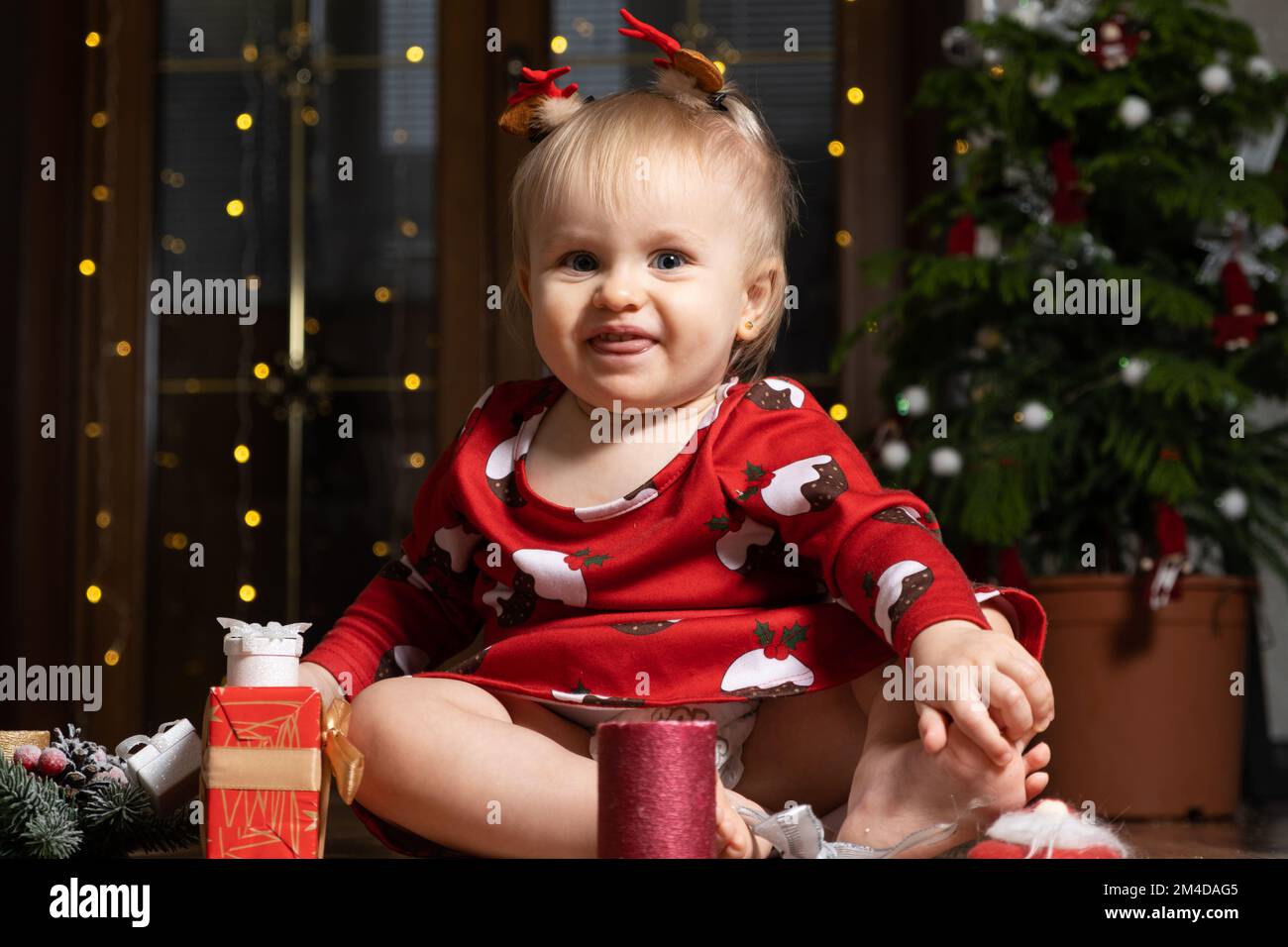 A one-year-old girl with blue eyes smiles sitting near a Christmas tree. Christmas, New Year and children. Stock Photo
