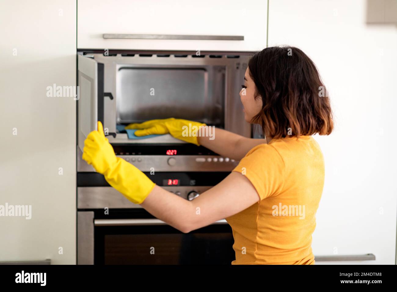 Happy Young Arab Woman Wiping Microwave With Rag While Cleaning In Kitchen Stock Photo