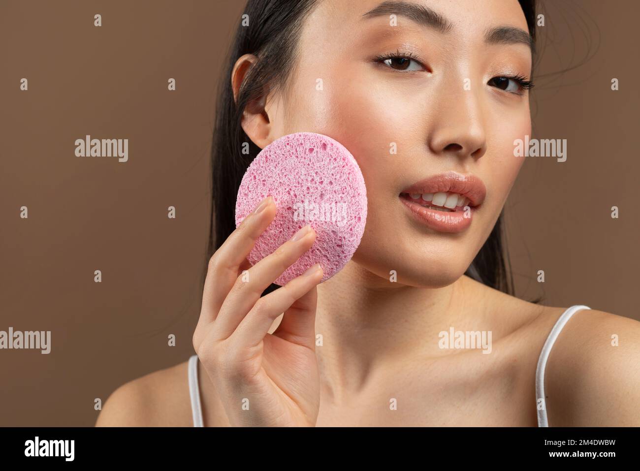 Face cleansing concept. Closeup of korean lady using soft facial cellulose sponge, washing face on brown background Stock Photo