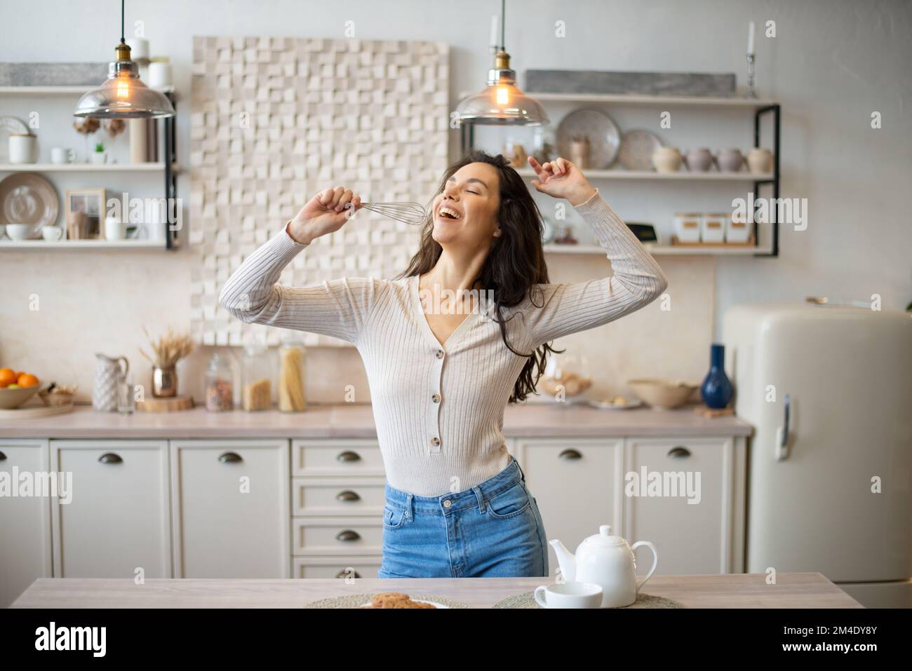 Playful young woman singing at appliance as imaginary microphone, dancing in light kitchen interior, copy space Stock Photo