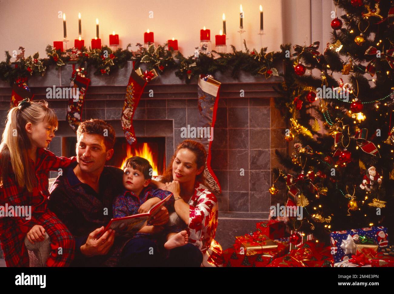 Young Caucasian family sitting on the floor in their pajamas in front of the Christmas tree and a roaring fire reading a Christmas book Stock Photo