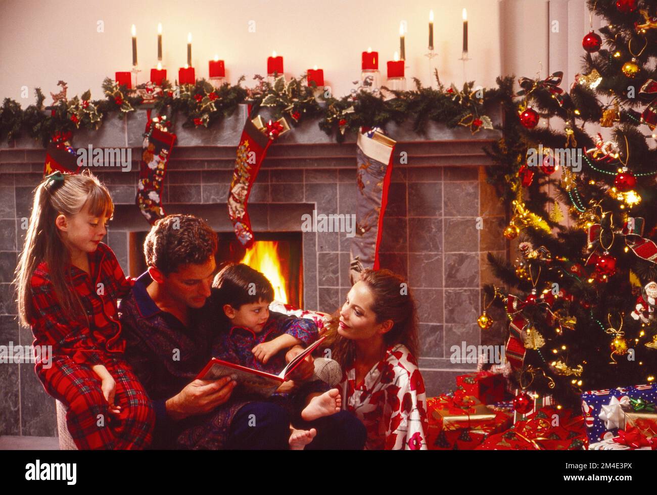 Young Caucasian family sitting in front of a Christmas tree and fireplace, all in their pajamas reading a book Stock Photo
