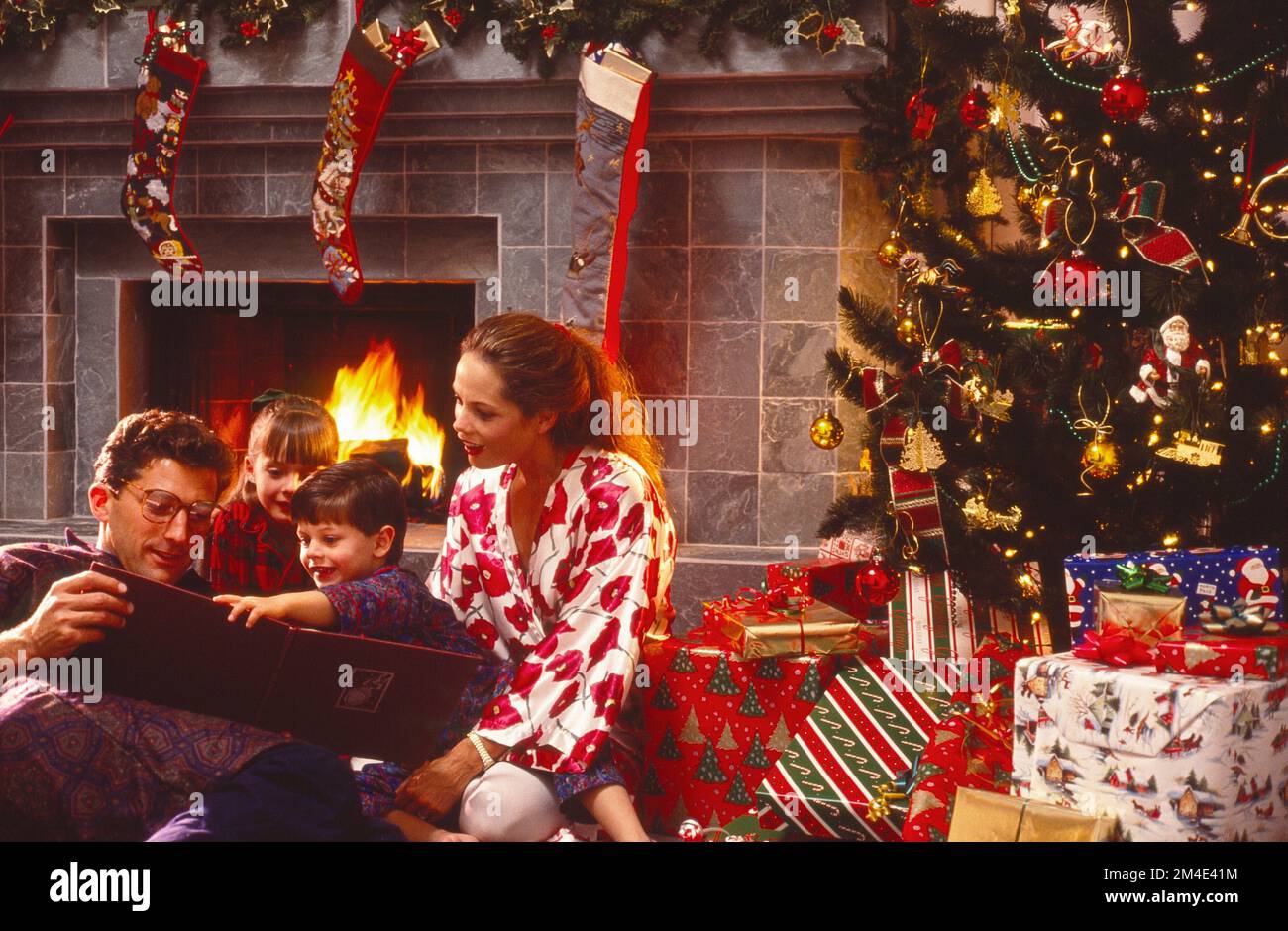 Family sitting on the floor in front of a fireplace with the Christmas tree presents and stockings hung from the mantel in the background Stock Photo