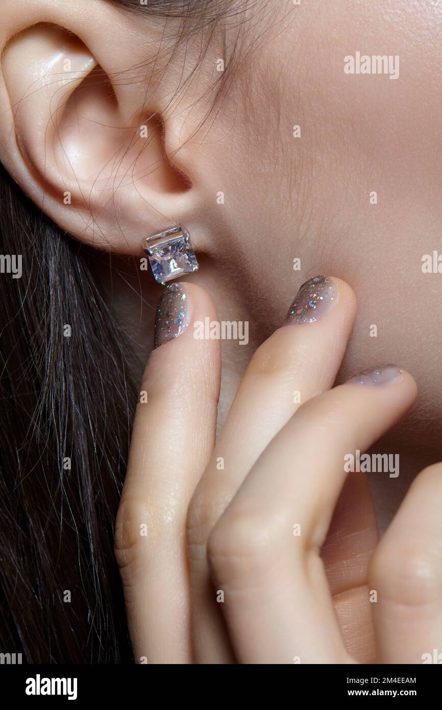Portrait of young woman on gray background. Female posing with hand near face. In the ear is an earring with a white crystal stone. Nails covered in g Stock Photo