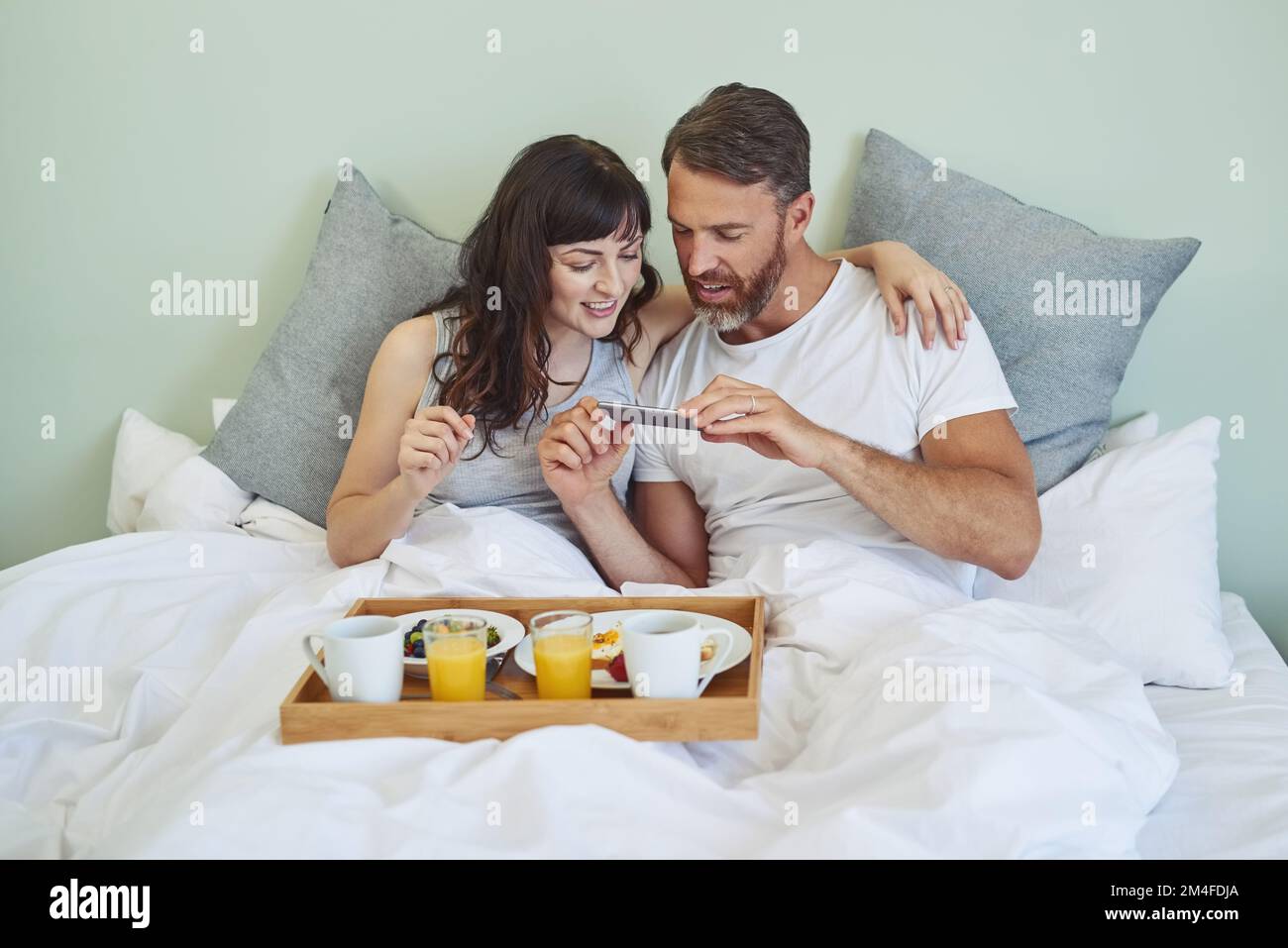 Lets make people jealous with a photo of our food. a cheerful young couple sitting in bed while enjoying breakfast together and taking a picture Stock Photo