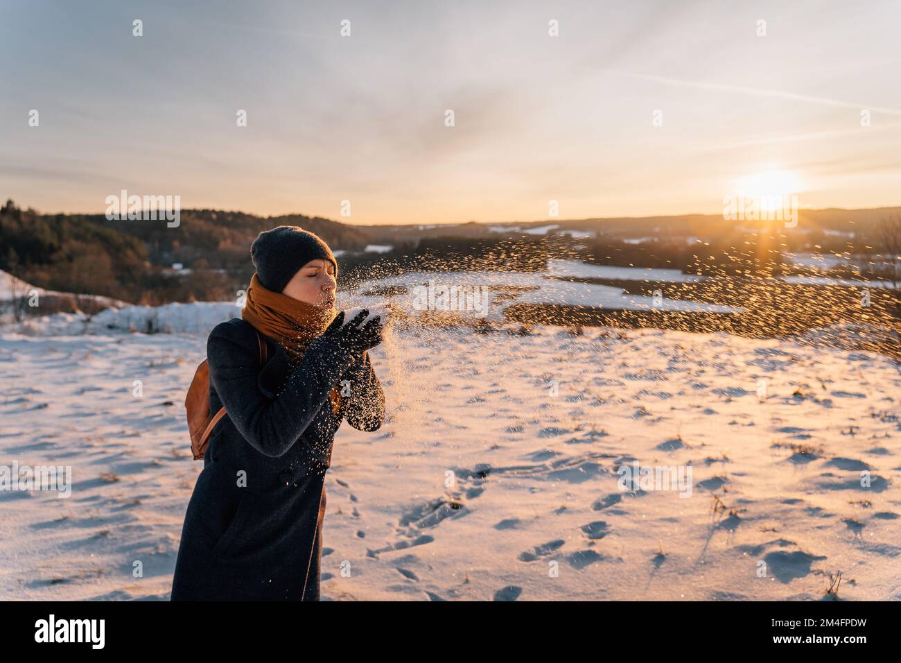 A woman blows in the palm of her hand with snow at sunset in winter Stock Photo
