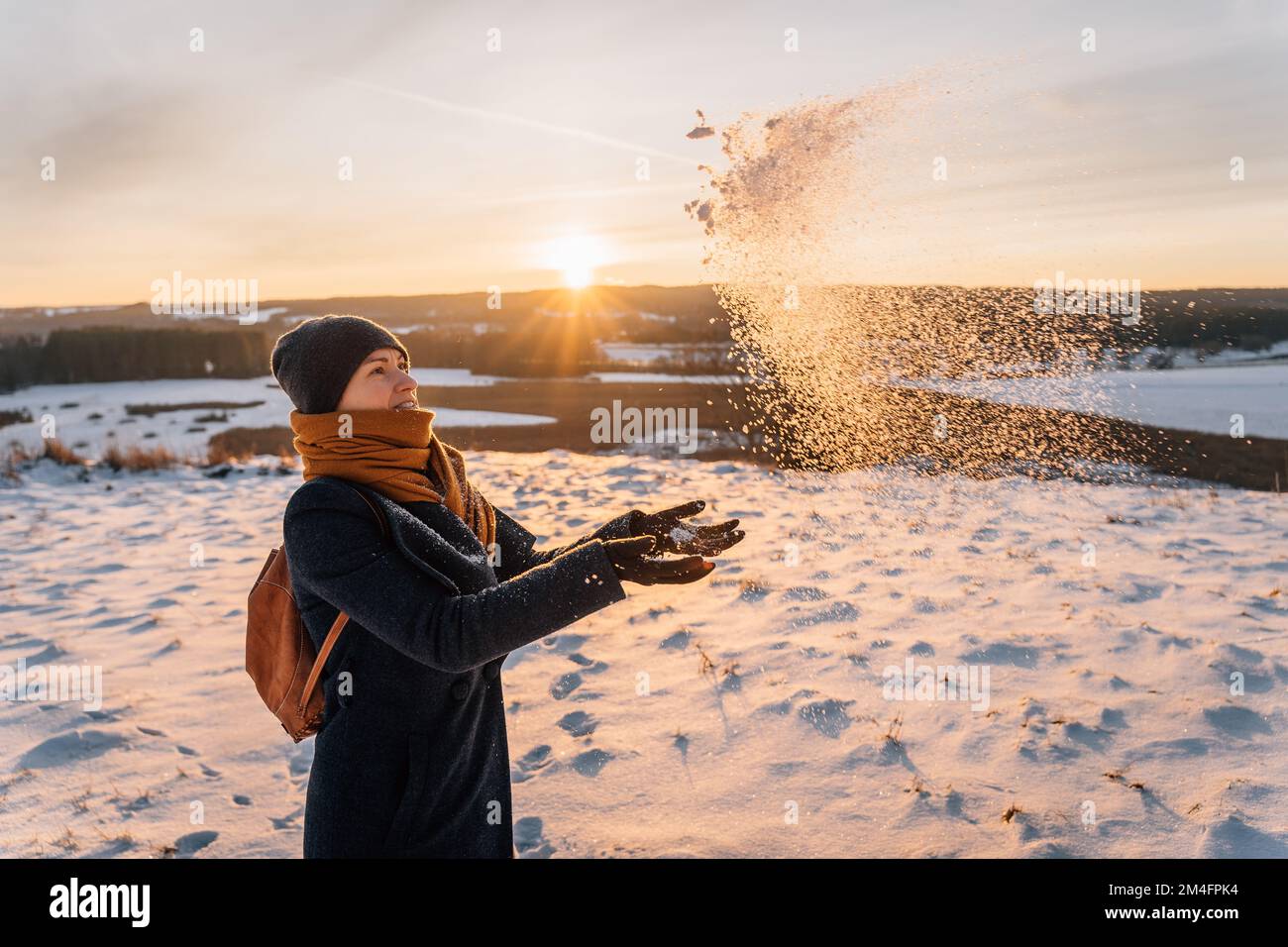 A woman in winter in warm clothes throws her palms and snow against the background of sunset Stock Photo