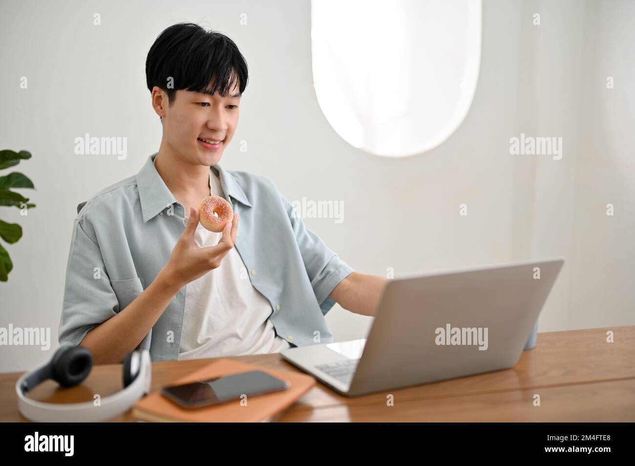 Handsome young Asian man enjoys eating donut while working on his ...