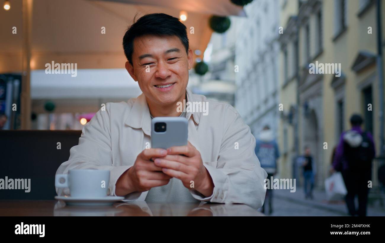 Smiling asian man tourist sitting in street cafe holding mobile phone booking tickets online remotely communicates with friends in social network on Stock Photo