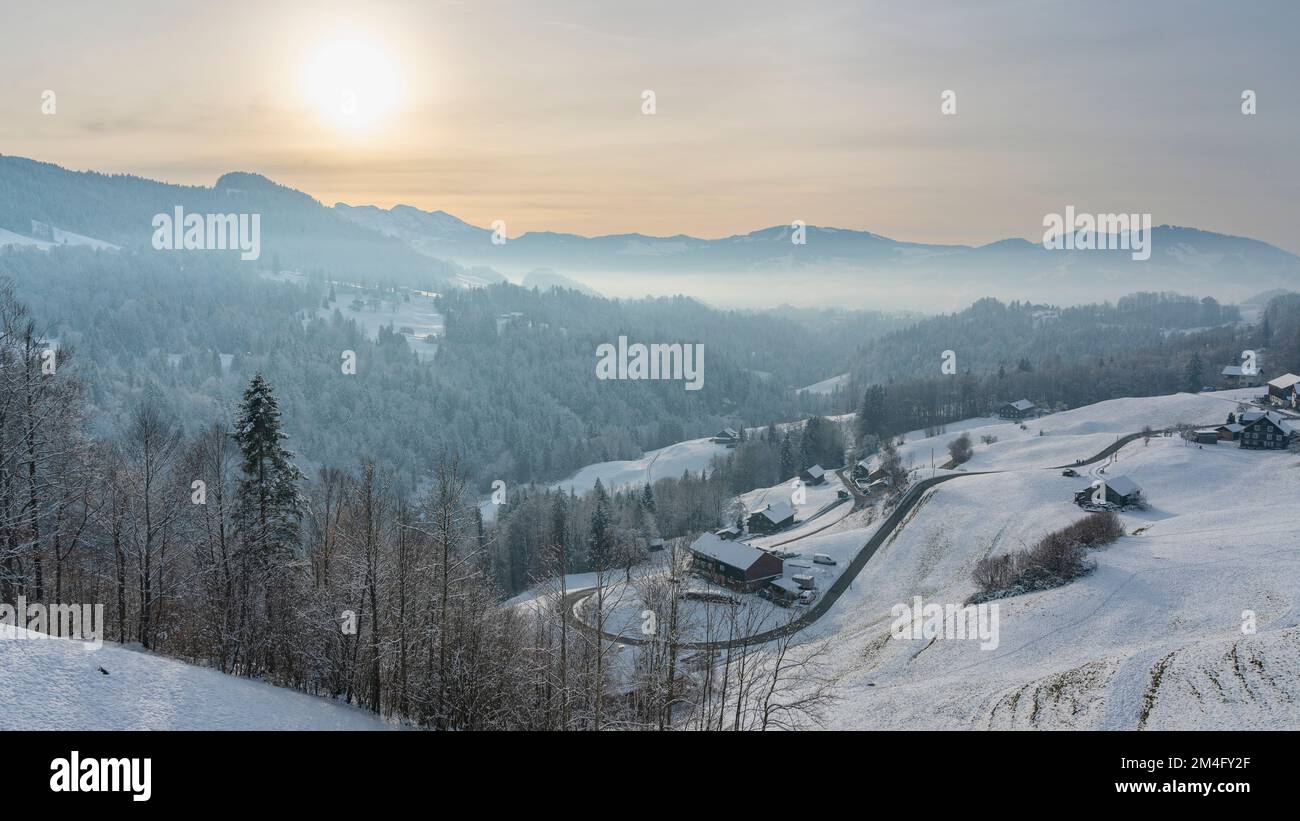 Frisch verschneite Bergstraße, die sich ins Tal schlängelt, Bauernhaus am Straßenrand, Berge und Täler mit Wäldern und Wiesen im Hintergrund, Abend Stock Photo