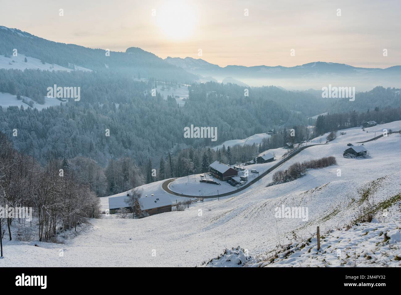 Frisch verschneite Bergstraße, die sich ins Tal schlängelt, Bauernhaus am Straßenrand, Berge und Täler mit Wäldern und Wiesen im Hintergrund, Abend Stock Photo