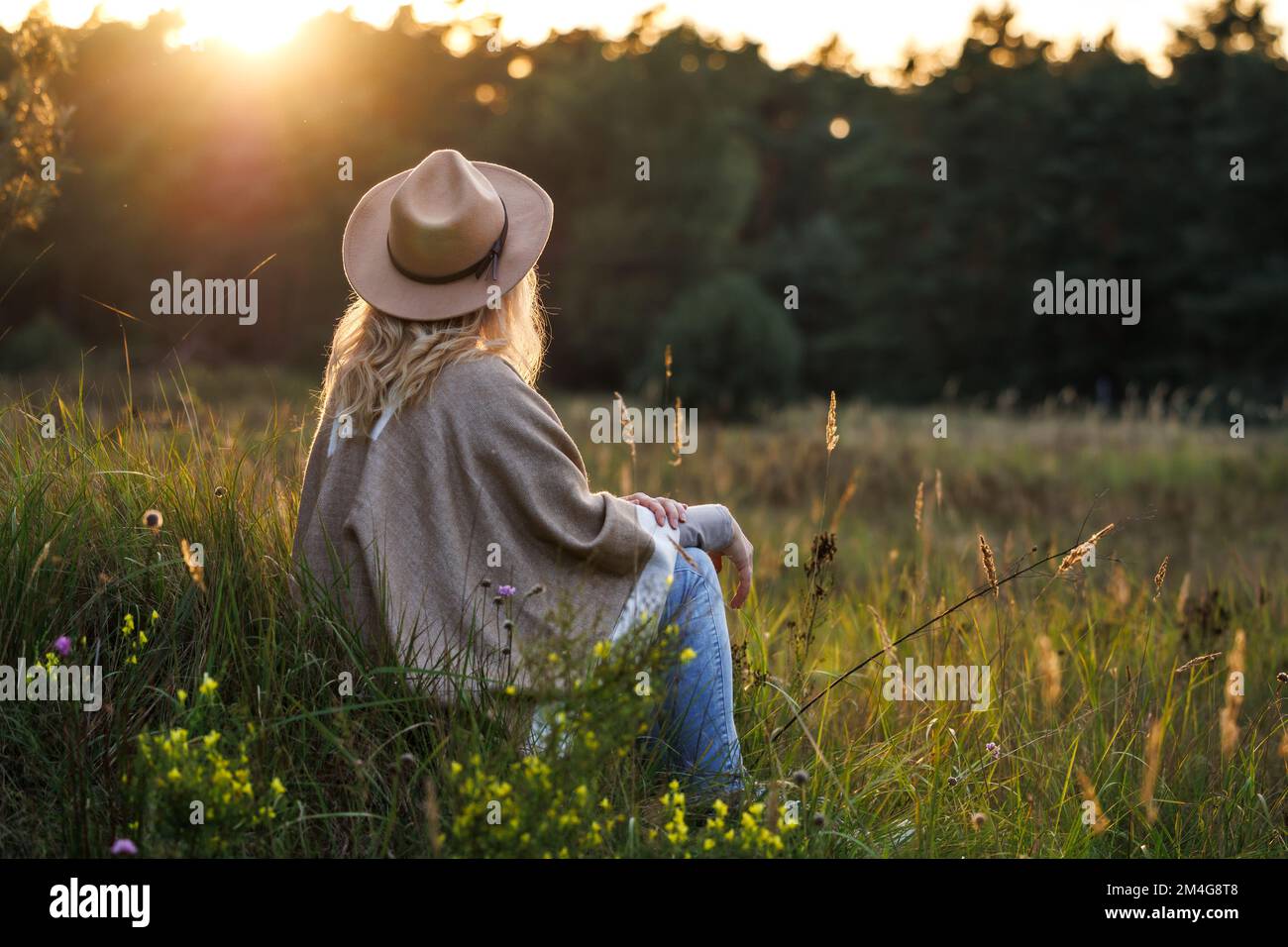 Relaxation outdoors at sunset. Boho woman with hat and poncho sitting in meadow. Enjoyment of indian summer in nature Stock Photo