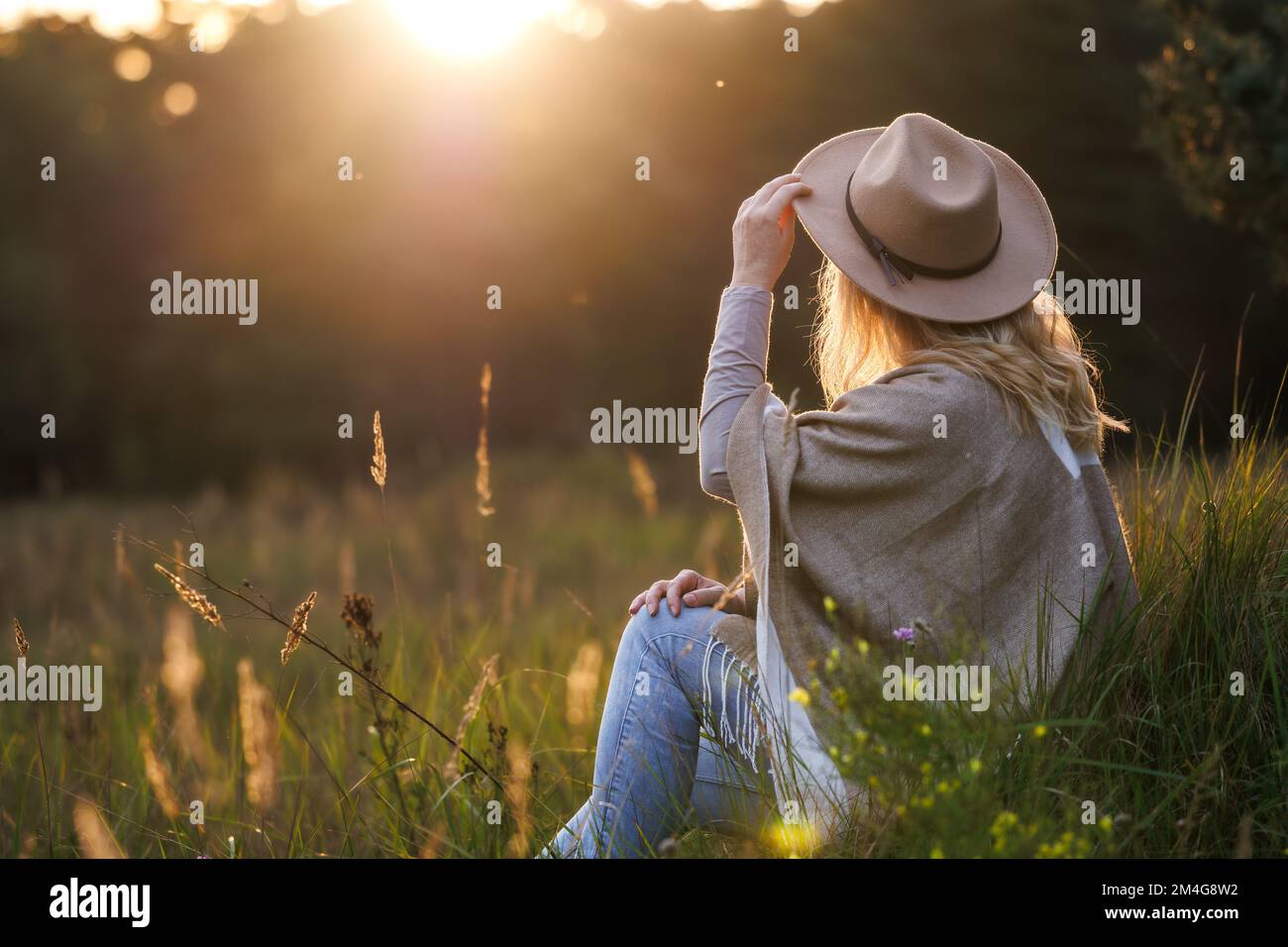 Happy woman with hat and poncho relaxing outdoors during sunset. Mental wellbeing and resting in nature Stock Photo