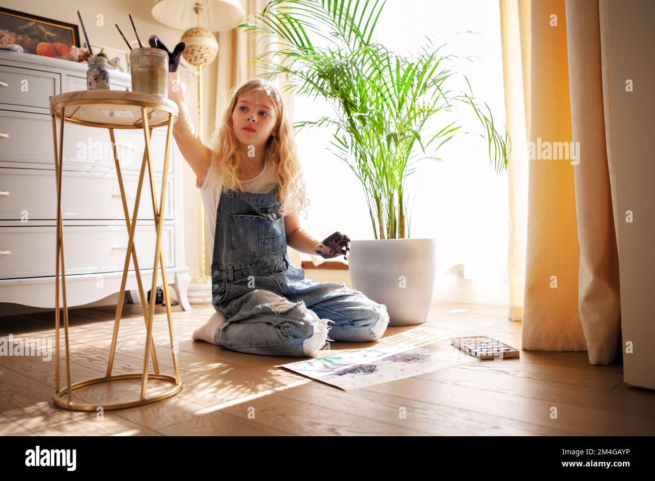 Child girl making drawings hands on paper sitting on floor. Young female artist painting with finger paints in sunny living room Stock Photo