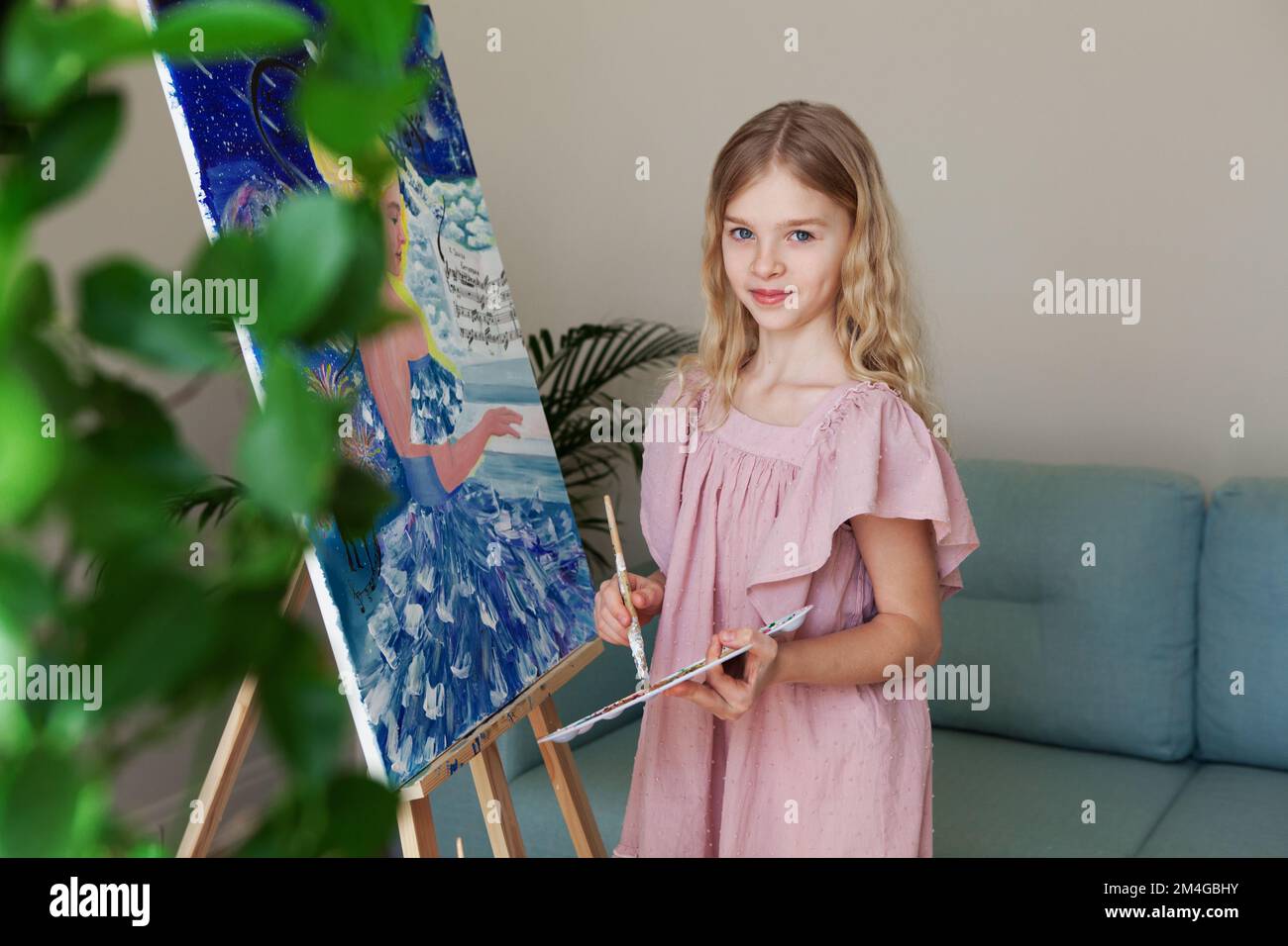 Blonde child girl artist drawing blue painting at home, looking in camera, wearing pink dress Stock Photo