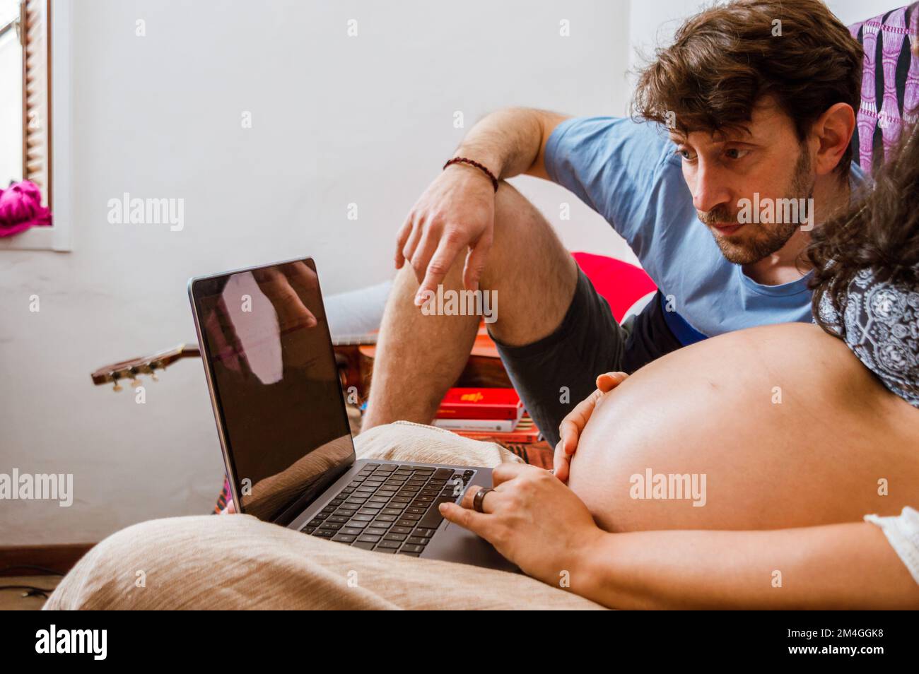 Caucasian man with beard, watching laptop screen with his pregnant wife while browsing the internet looking for prices of baby products, sitting on th Stock Photo