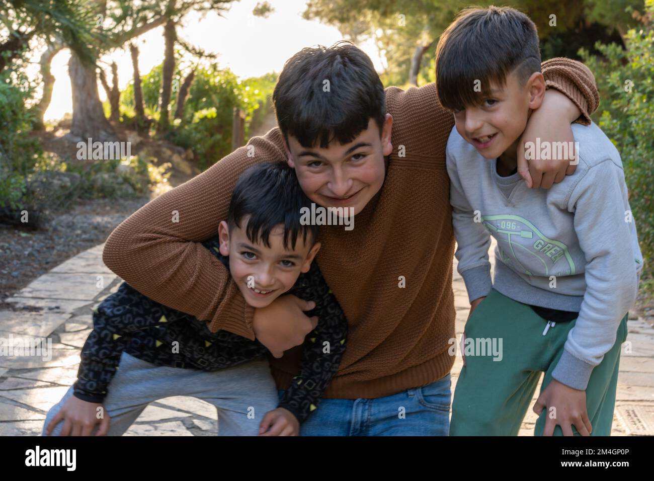 Happy teenager in casual clothes smiling and embracing younger brother while having fun on paved path in evening in park Stock Photo