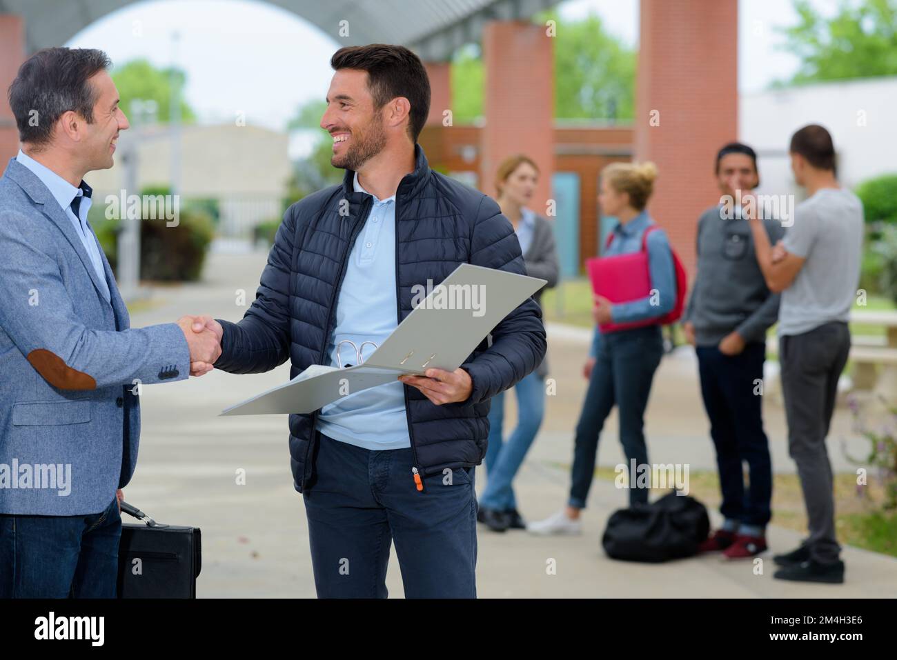 two mature men shaking hands on campus students in background Stock Photo