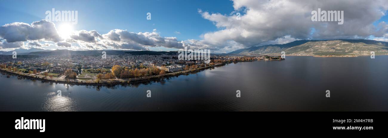 Greece, Giannena city Pamvotida Lake, Epirus. Drone, aerial panoramic view of Ioannina, sun behind cloudy blue sky background makes sparkle water. Ban Stock Photo