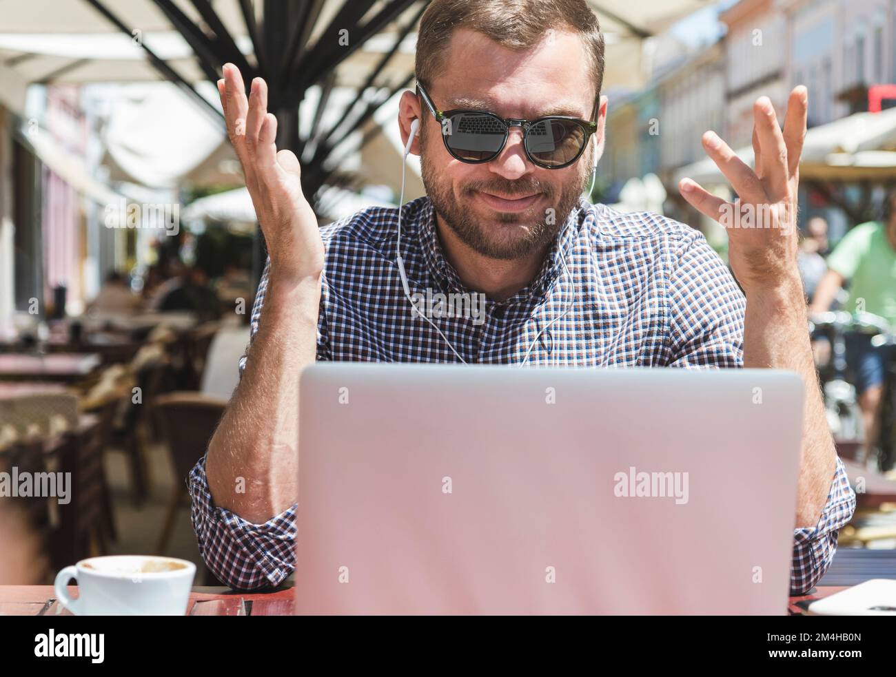 Handsome adult man in sunglasses talking video call using his headphones and laptop while sitting in an outdoors cafe. Stock Photo