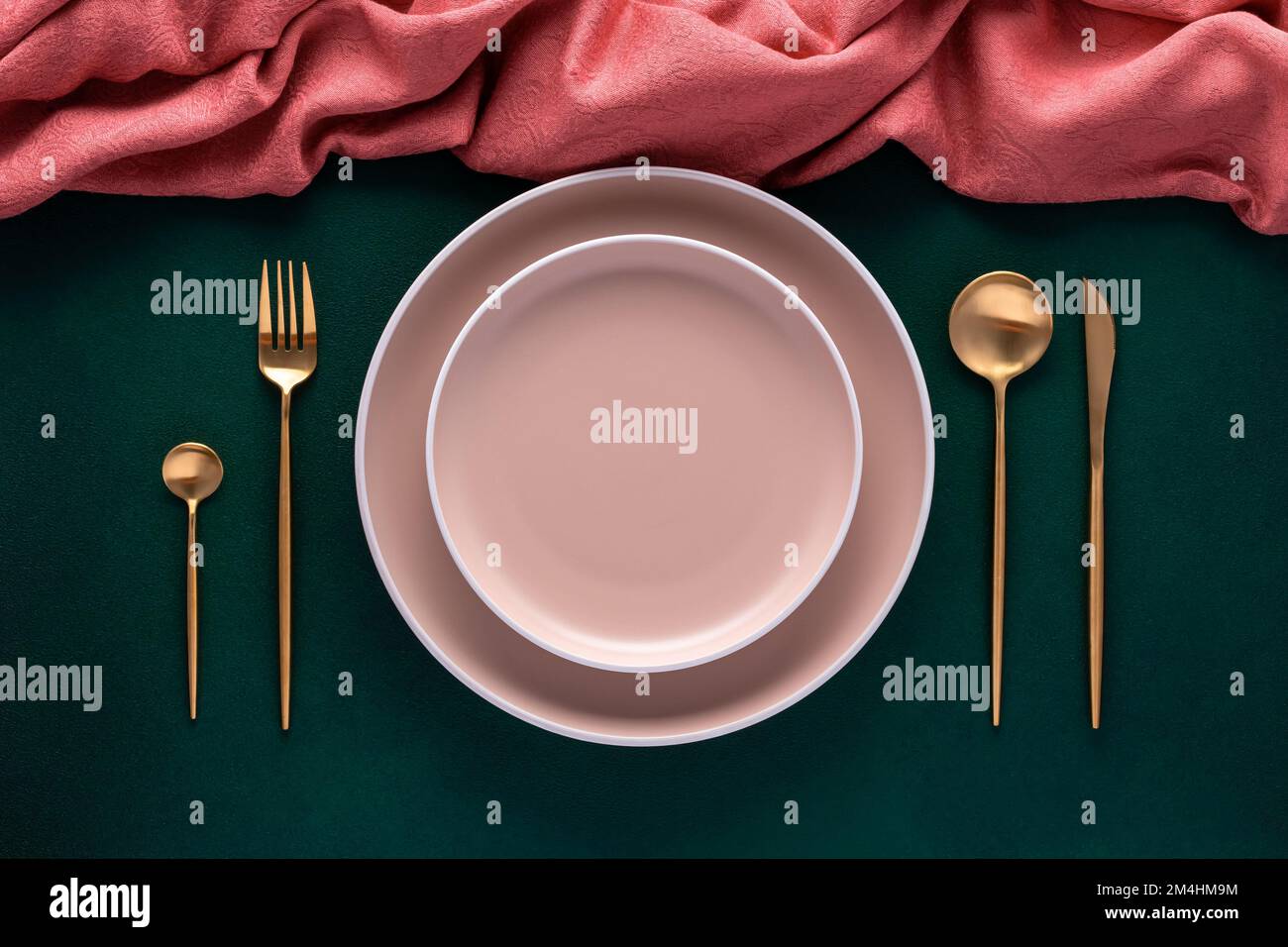 Festive place setting with pink napkin. Empty plates and gold cutlery on dark green background. Top view. Dinner table in luxury restaurant. Card or m Stock Photo