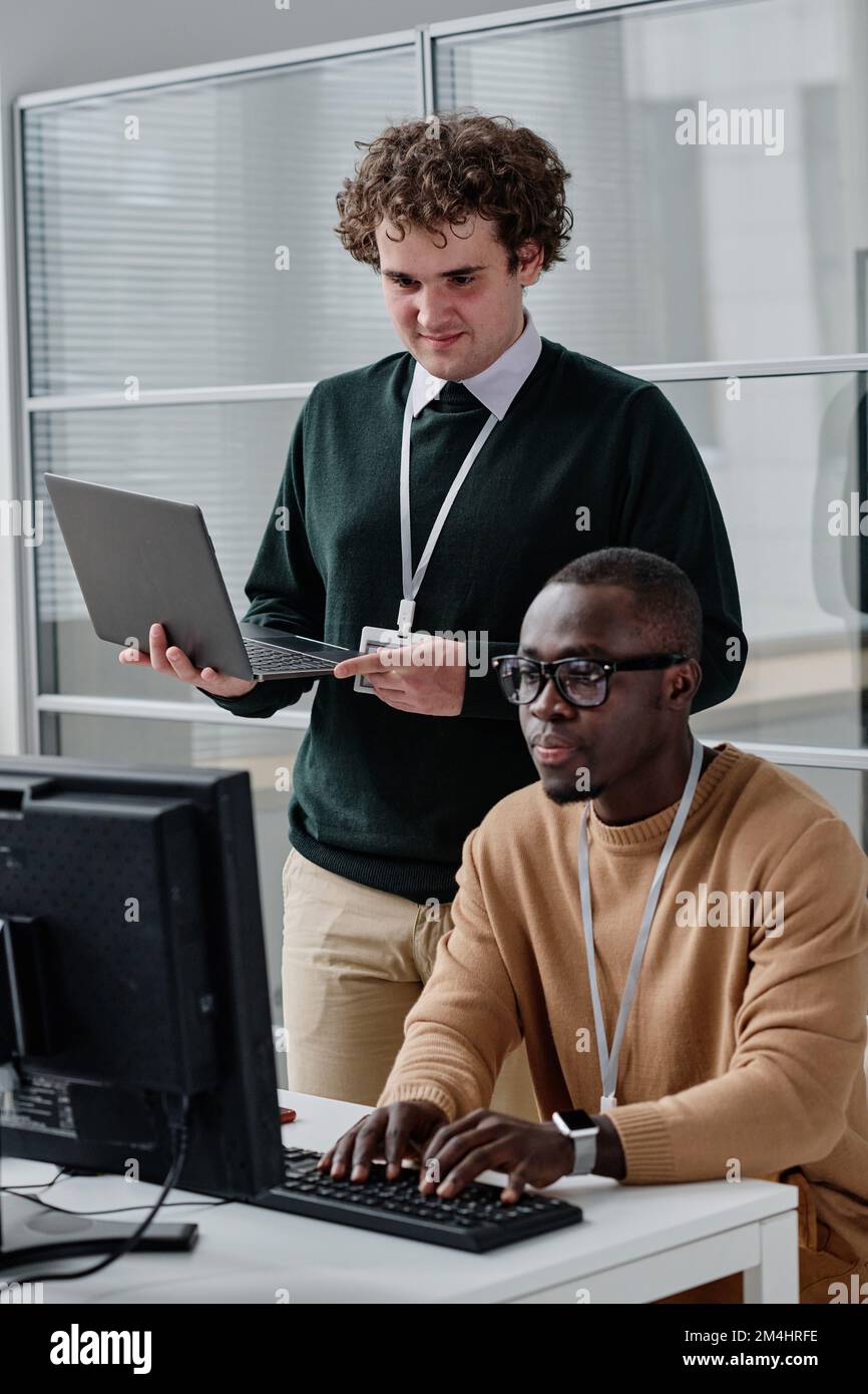 African American developer typing codes on keyboard with his colleague standing in background and checking codes on his laptop Stock Photo