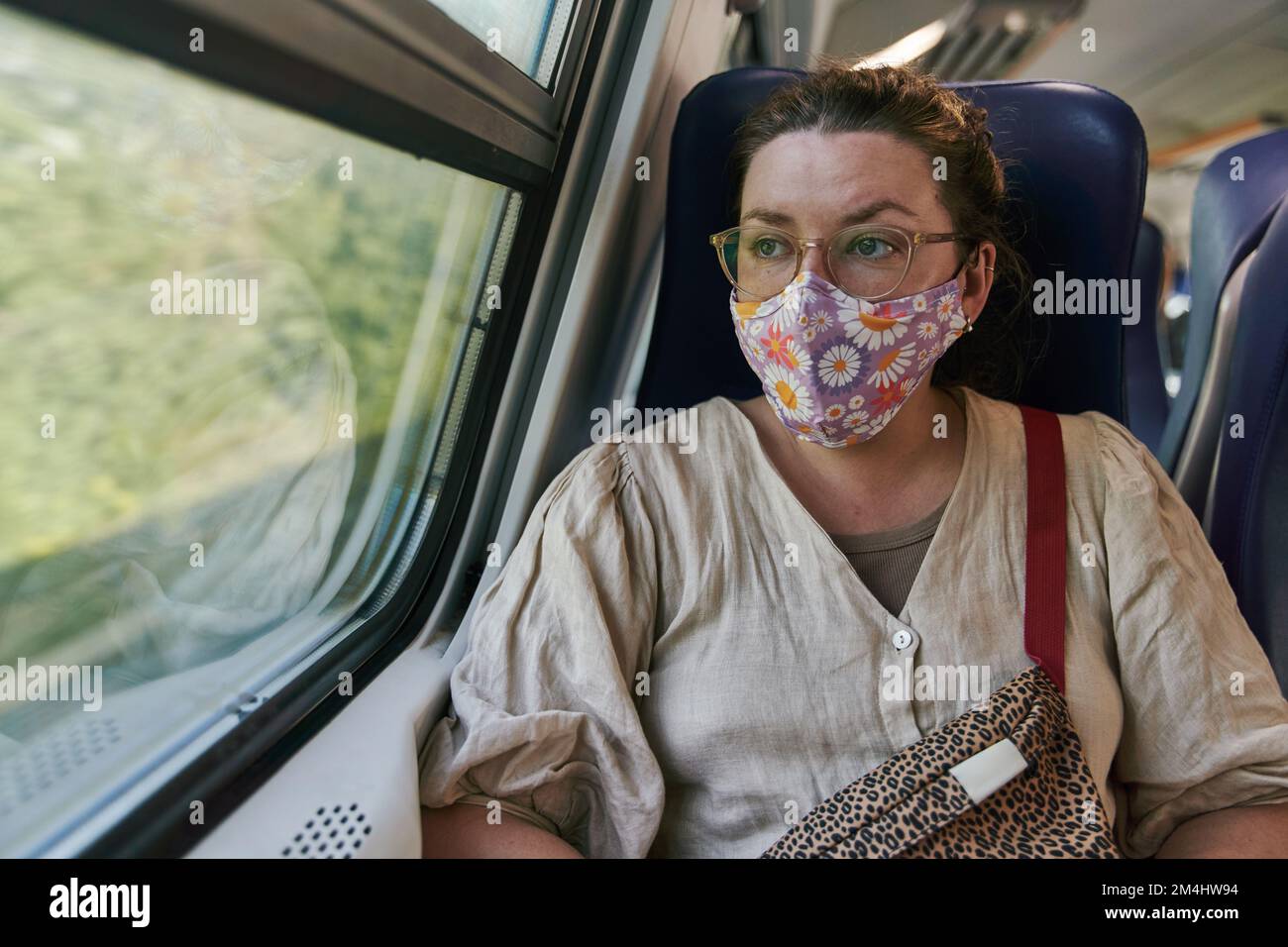 A girl in glasses and a medical mask riding a train and looking out the window Stock Photo