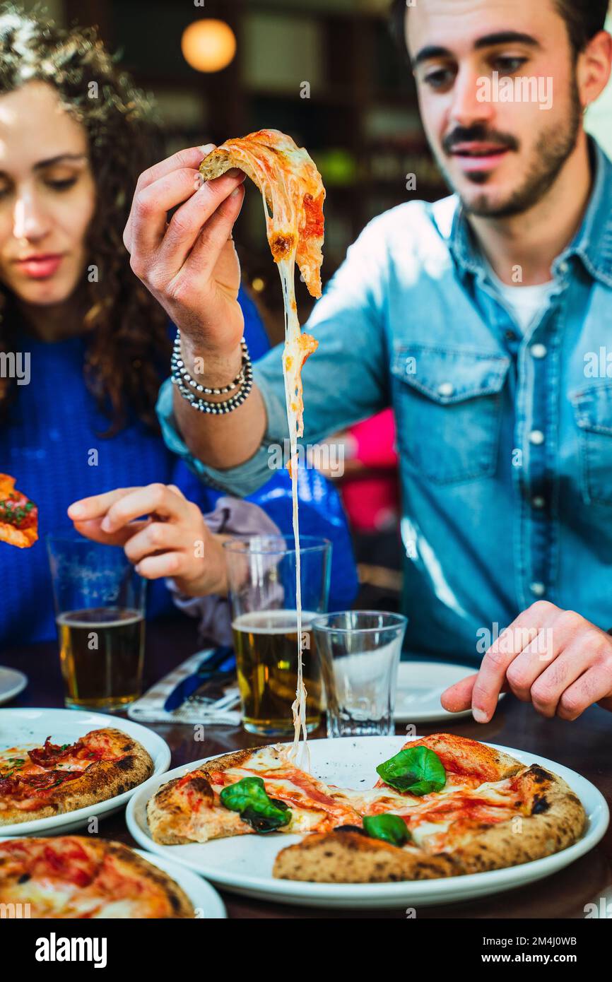 Guy taking a pizza slice out from the plate. The cheese stretching and looking appetizing. Friends enjoying cheese pizza with crispy crust. Beer glass Stock Photo