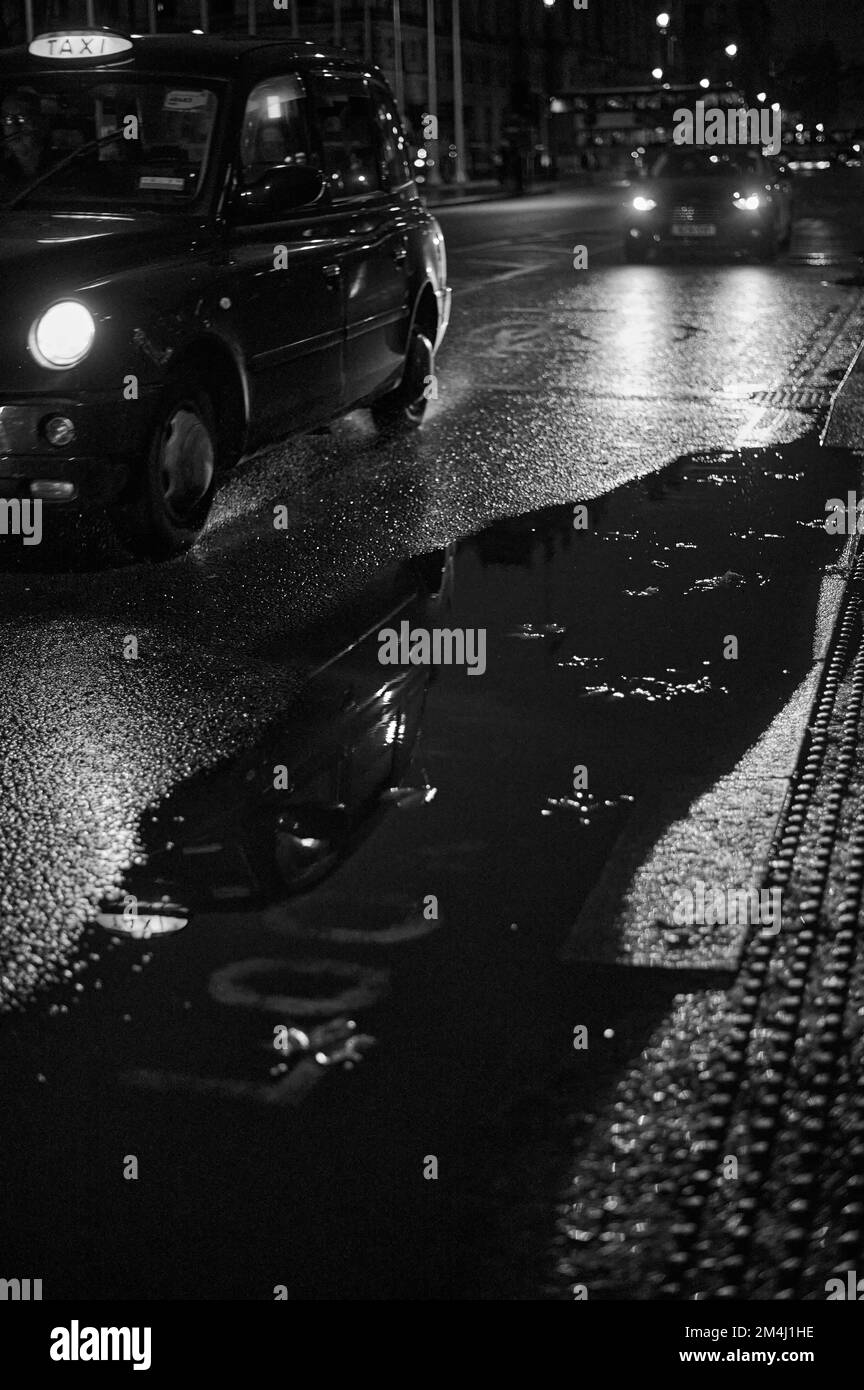 Black and white monochrome image of blurred background with London black cab on wet road with large puddle Stock Photo