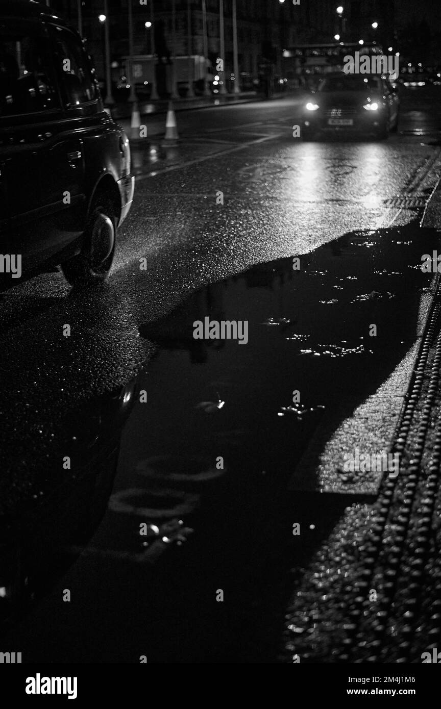 Black and white monochrome image of blurred background with London black cab on wet road with large puddle Stock Photo