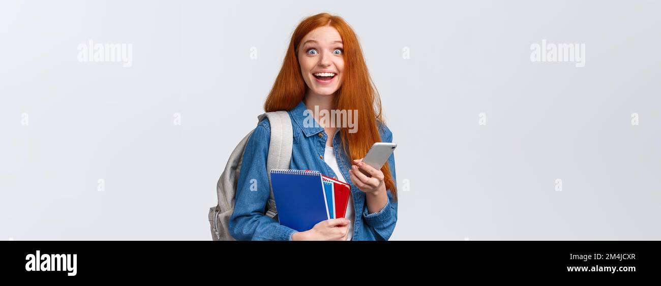 Excited and happy, relieved cute redhead female college student with backpack, notes in notebook and smartphone, looking cheerful camera, texting Stock Photo