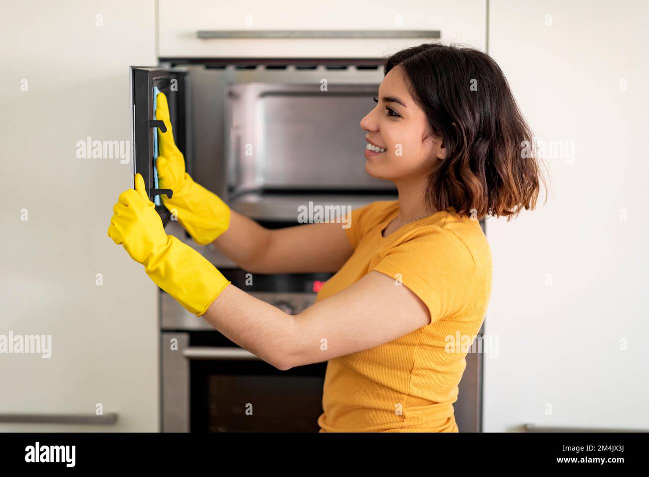 Housekeeping Concept. Smiling Arab Woman In Rubber Gloves Wiping Microwave From Dirt Stock Photo