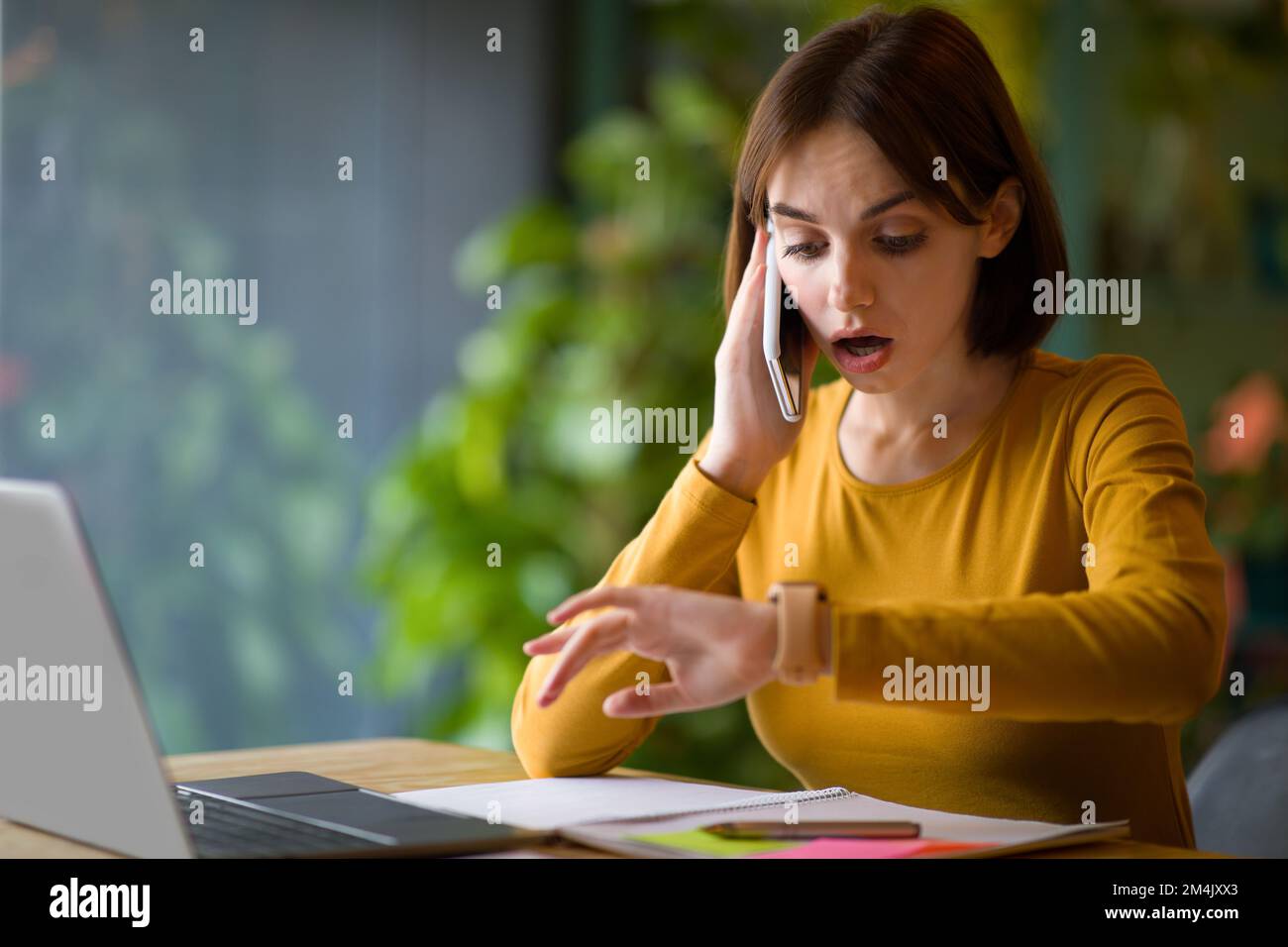 Overworked young woman checking time while having phone conversation Stock Photo