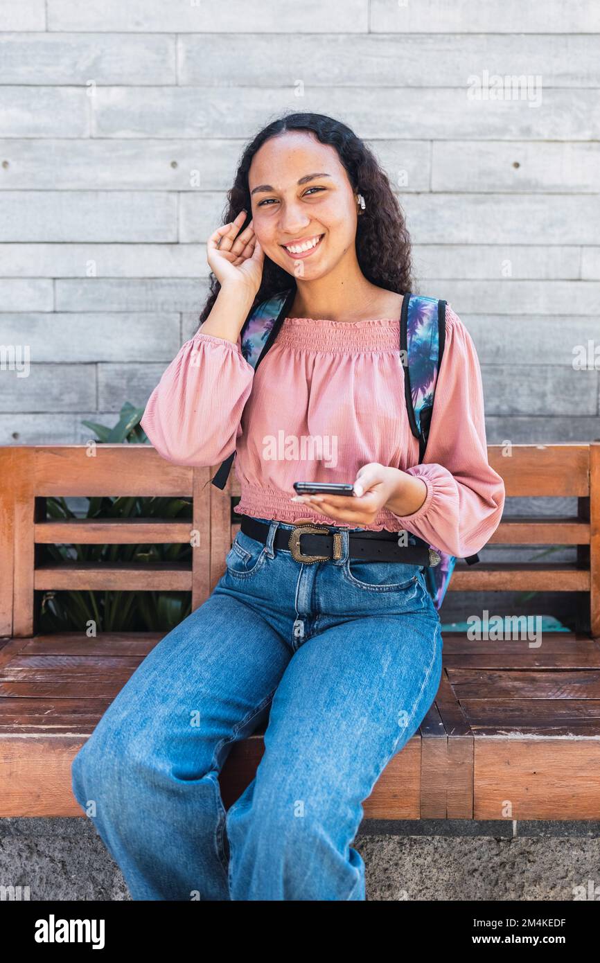 Latin university student woman smiling and using her mobile sitting outside the mall in her free time Stock Photo