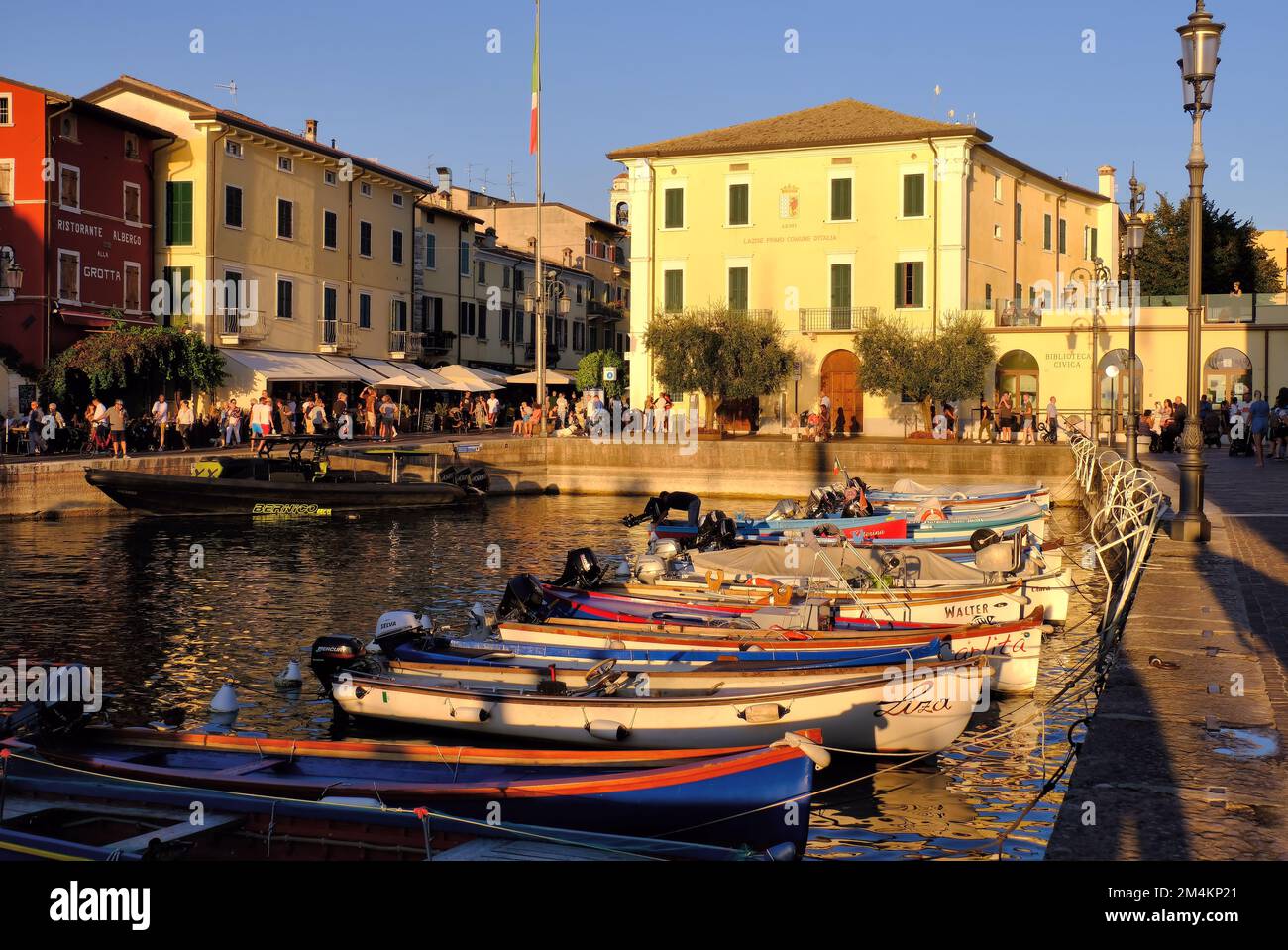 Lazise: Fishing port and marina with boats moored and restaurants glowing gold soon before sunset at Lazise on Lake (Lago) Garda, Veneto, Italy Stock Photo