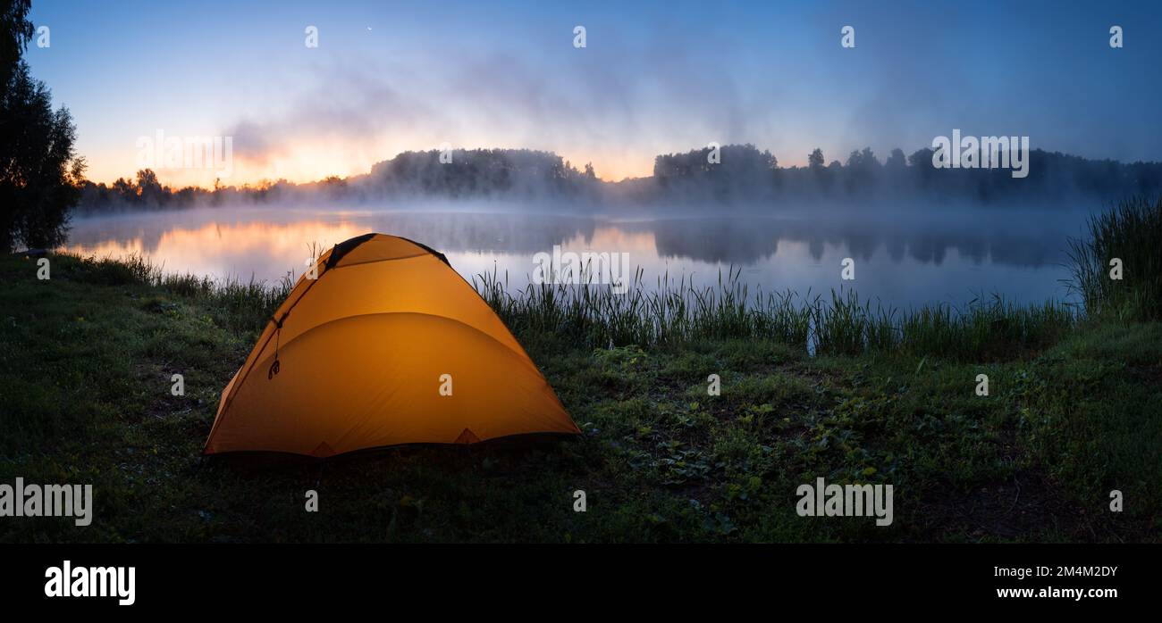 Orange tent lit from inside on bank of foggy river in early morning Stock Photo