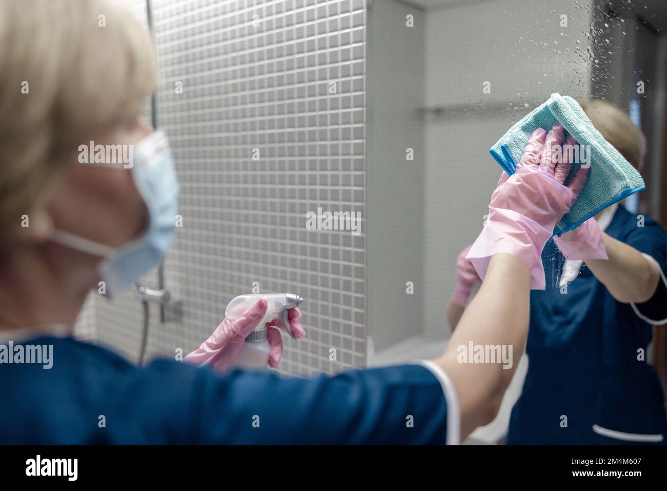 Chambermaid in mask cleans mirror in hotel bathroom spraying detergent on surface Stock Photo