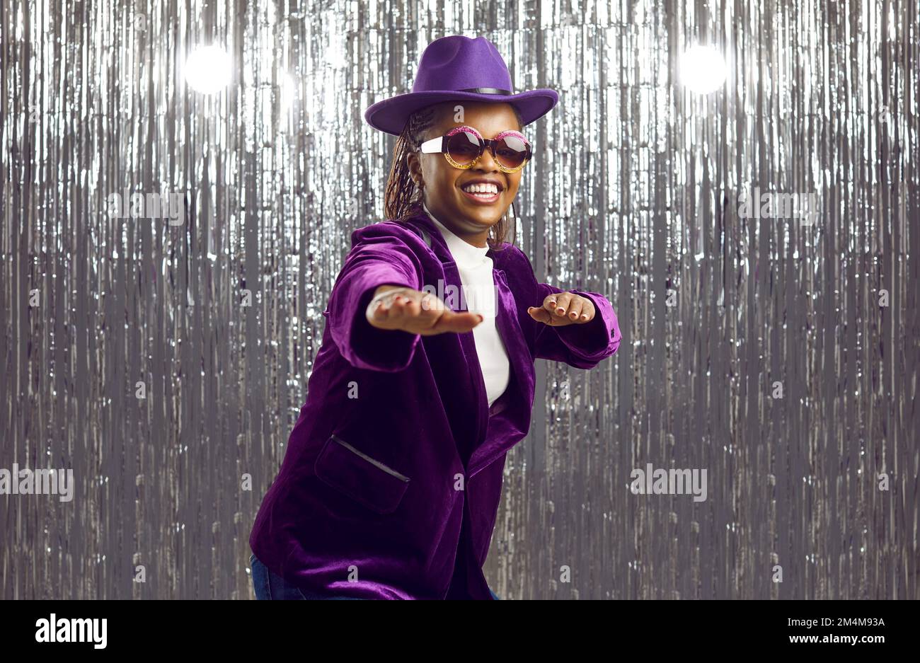 Happy young Black girl in purple hat, jacket and glasses dancing and having fun at disco party Stock Photo