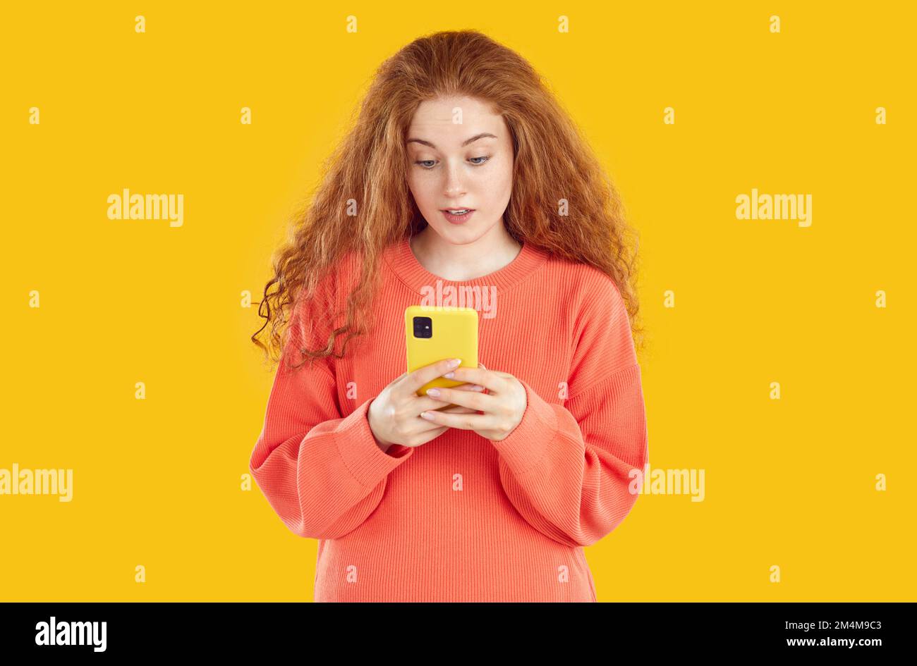 Frustrated amazed redhead girl with curly hair looking at smartphone screen on yellow background. Stock Photo