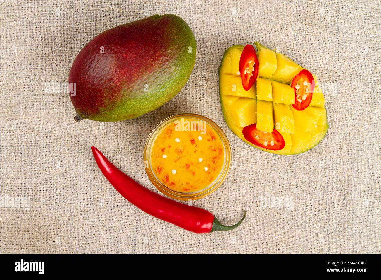 Top view of Whole mango, whole chili pepper, sliced hot pepper, cutted half of mango and Spicy mango chutney in glass bowl on burlap. Stock Photo