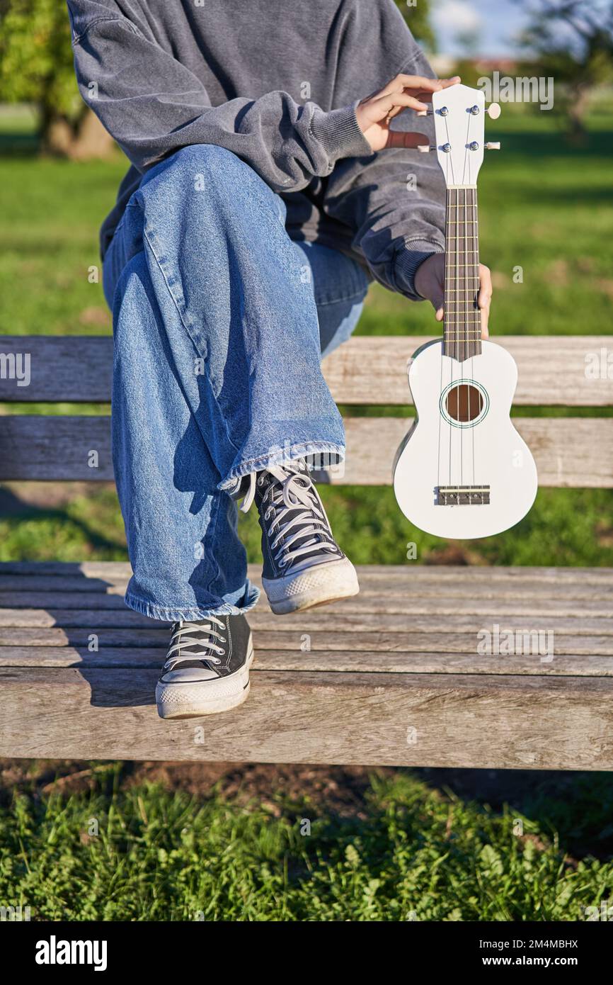cropped shot of young girl in sneakers and jeans hands holding ukulele musical instrument while she sits on bench in green sunny park 2M4MBHX