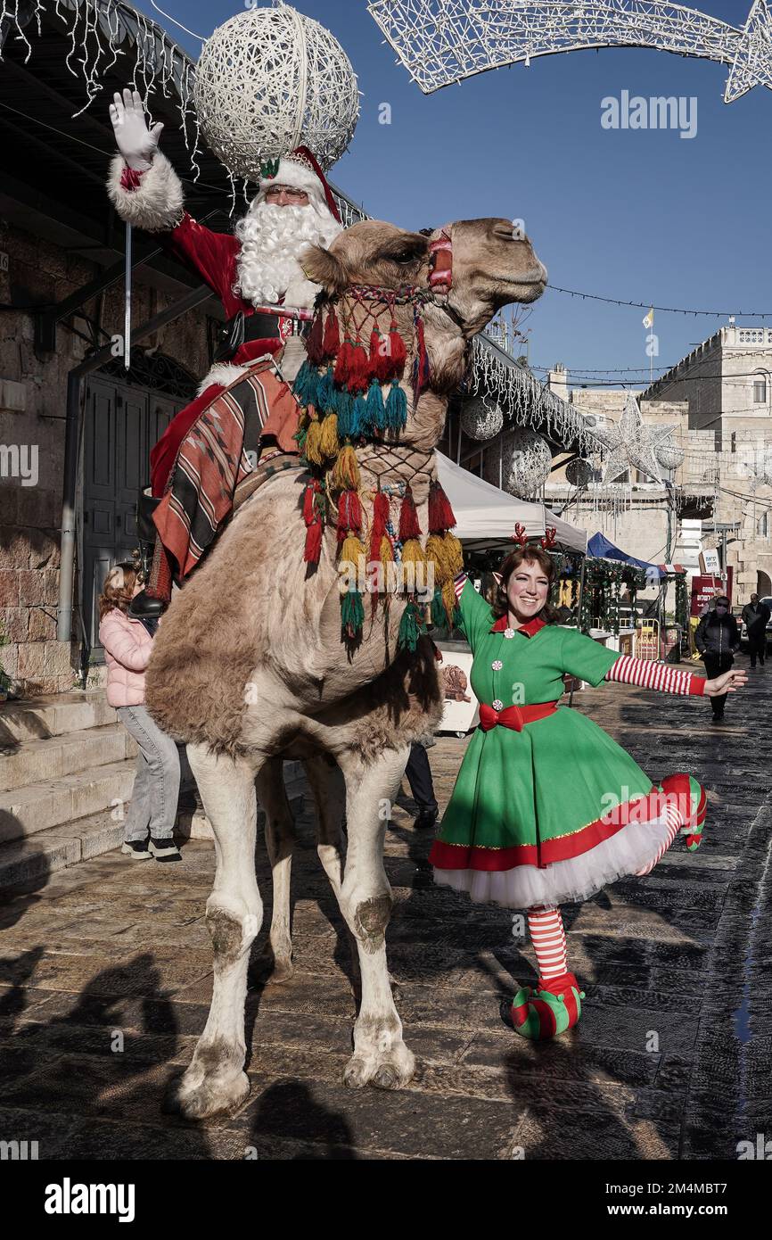 Jerusalem, Israel. 22nd Dec, 2022. Santa Claus, or 'Baba Noel' as he is called in Arabic, is escorted by an elf as he rides a camel substitute for reindeer at Jerusalem's Old City New Gate. The Jerusalem Municipality and the Jewish National Fund distributed specially grown Arizona Cypress Christmas trees to the Christian population at the Jaffa Gate. Credit: Nir Alon/Alamy Live News Stock Photo