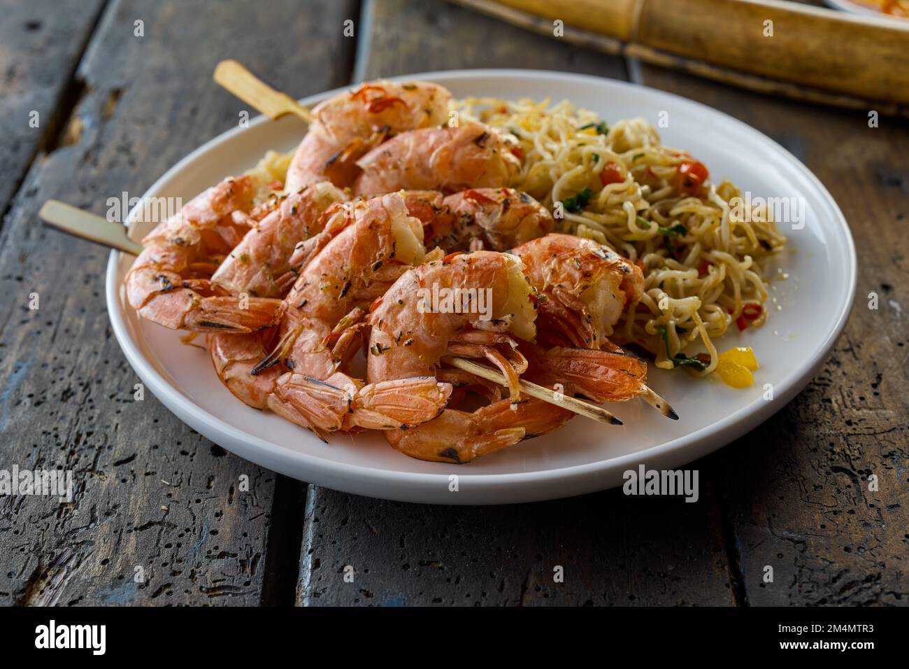 Appetizing Asian shrimps scampies on skewers with tasty noodles served on white ceramic plate on wooden table in light restaurant Stock Photo