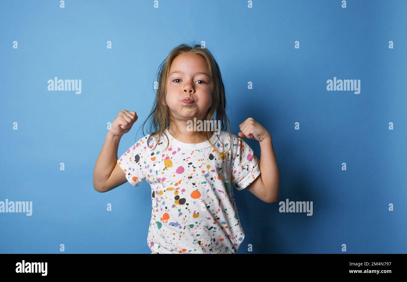 a little girl shows strength by raising her arms up and puffing out her cheeks on an isolated blue background Stock Photo