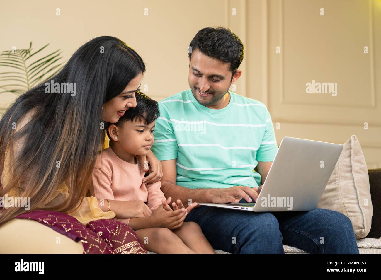 focus on kid, happy couple with sad kid using on laptop on sofa at home - concept of checking school admission or selection, seeing toys and ecommerce Stock Photo