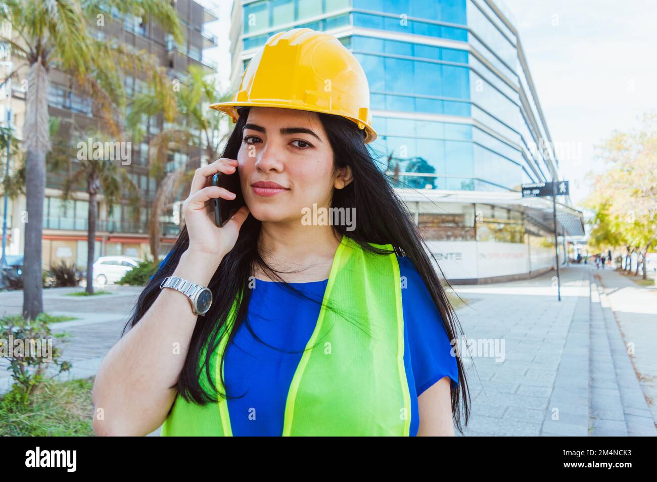 young caucasian venezuelan female engineer, wearing protective helmet and safety vest, standing outdoors talking on the phone and looking at the camer Stock Photo