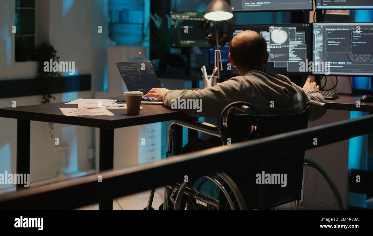 System engineer with impairment working on web programming, using laptop and multiple screens. Male IT coder with chronic disability in wheelchair developing new user interface. Stock Photo