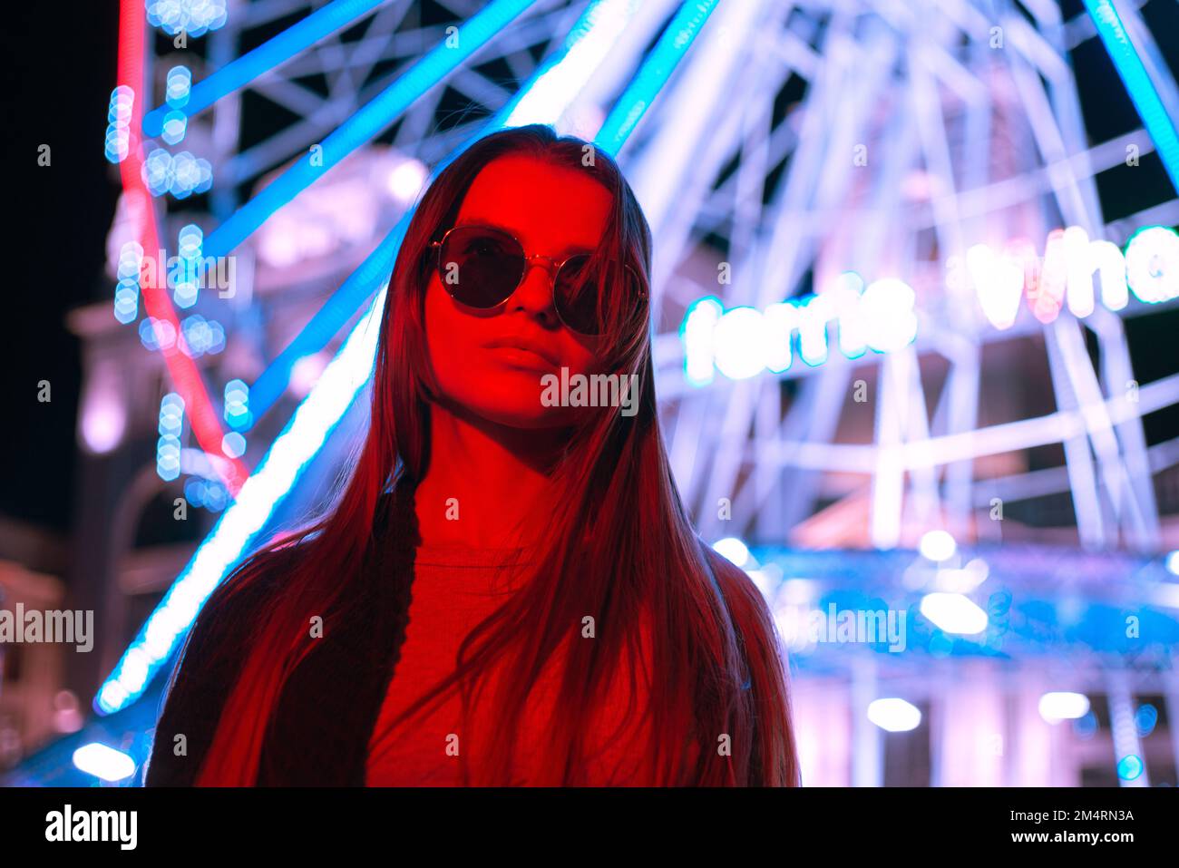 Portrait a pretty tourist urban young woman in sunglasses standing In front of a Ferris wheel in a red neon light-colored background Stock Photo
